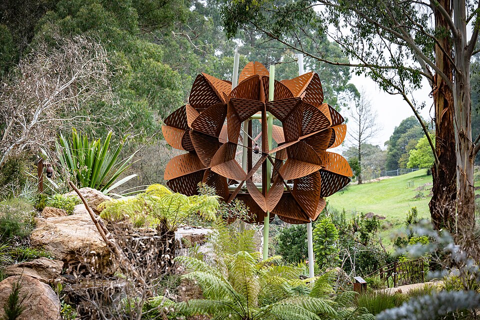 A rust-coloured metal sculpture among rocks, ferns and trees in a garden.