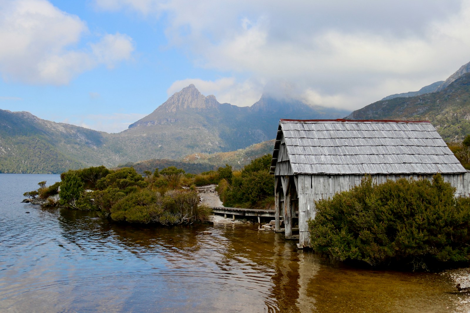 A weathered timber boatshed at the edge of Dove Lake, with Cradle Mountain rising behind it.